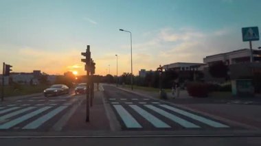 Gün batımında ağaç siluetleri ve modern endüstriyel ya da okul binası olan huzurlu bir banliyö manzarası. Sıcak akşam ışığı şehrin kenarında sakin ve dengesiz bir atmosfer yaratıyor..