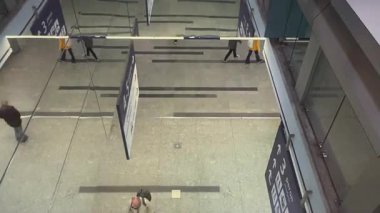 Modern bir kamu binasında, muhtemelen bir havaalanı, tren istasyonu ya da kongre merkezinde, geniş bir salonda hareket eden insanların genel görünümü. Yansıtıcı yüzeyler ve işaretler teknolojiden anladığını gösteriyor.