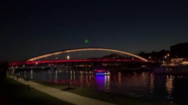 Polonya 'nın Krakow kentindeki Vistula Nehri üzerindeki Bernatka Footbridge' in sakin bir akşam görüntüsü. Sokak lambaları suyu yansıtıyor ve alacakaranlıkta huzurlu bir şehir atmosferi yaratıyor..