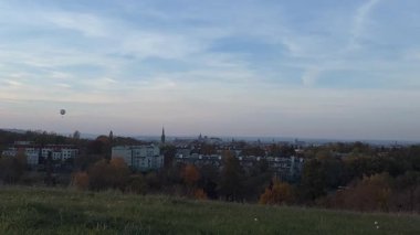 Krakow, Polonya 'nın panoramik manzarası, gün batımında çimenli bir tepeden, sıcak hava balonuyla birlikte. Şehrin cazibesini ve açık gökyüzünün güzelliğini yansıtan huzurlu bir an..