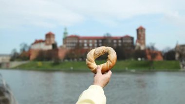 Geleneksel bir Krakow simit obwarzanek 'i tarihi Wawel Kalesi ve Vistula Nehri' nin arka planına karşı tutarak. Polonya 'da yerel yiyecek, kültür ve seyahatin mükemmel bir kombinasyonu.