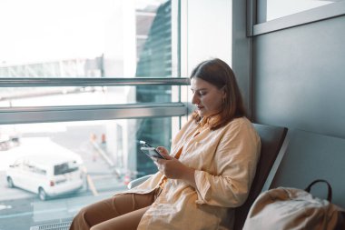 Havaalanı lobisinde rahat sarı elbiseli genç bir gezgin uçuşunu beklerken iki cep telefonu kullanıyor. Modern seyahat, teknoloji ve boş zamanlarla ilgili konular için mükemmel..