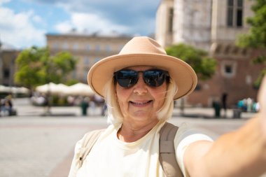 Yaşlı kadın şehir meydanında selfie çekerken gülümsüyor, elini sallıyor. Bej bir şapka ve güneş gözlüğü takıyor, güneşli bir günde etrafı geziyor..