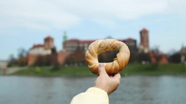 Geleneksel bir Krakow simit obwarzanek 'i tarihi Wawel Kalesi ve Vistula Nehri' nin arka planına karşı tutarak. Polonya 'da yerel yiyecek, kültür ve seyahatin mükemmel bir kombinasyonu.