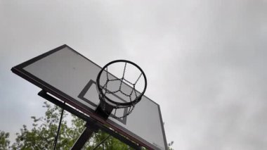 Ağaç dalları ve bulutlu gökyüzü tarafından çerçevelenmiş eski bir basketbol potası ve arka tahtanın alçak açılı görüntüsü. Kentsel spor ortamı gençlik kültürü, motivasyon ve aktif yaşam tarzı görselleri için mükemmel..