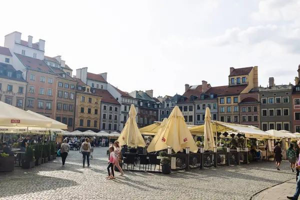 Polonya 'nın Varşova kentindeki Eski Kasaba Meydanı' nın geniş bir manzarası var. Arka planda açık kafeleri ve tarihi renkli binaları kaplayan sarı şemsiyeler var..