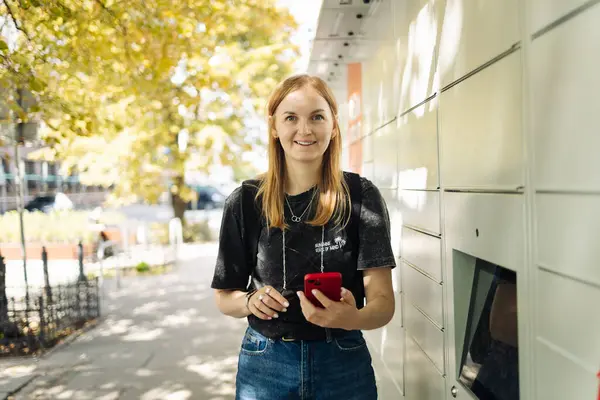Genç bir kadın güneşli bir günde açık havada bir dolap kullanıyor. Siyah tişörtlü ve kot pantolonlu sıradan bir sarışın kadın elinde akıllı bir telefon, self servis paket terminalinin yanında duruyor. Modern kavram