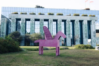Metalden kanatları olan pembe stilize edilmiş bir atın yer aldığı çağdaş açık hava sanat kurulumu. Geometrik heykel, kolonları olan modern cam bir binanın önündeki çimlerde sergileniyor.. 