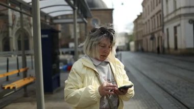 Polonya, Krakow 'da bir tramvay istasyonunda beklerken elinde akıllı telefon tutan kıdemli bir kadın. Bir Avrupa şehrinde kentsel hareketlilik, teknoloji kullanımı ve bağımsız yaşam gösteren modern bir seyahat anı.