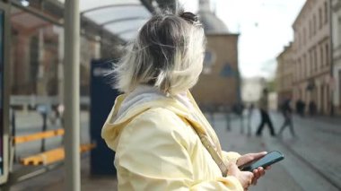 Polonya, Krakow 'da bir tramvay istasyonunda beklerken elinde akıllı telefon tutan kıdemli bir kadın. Bir Avrupa şehrinde kentsel hareketlilik, teknoloji kullanımı ve bağımsız yaşam gösteren modern bir seyahat anı.