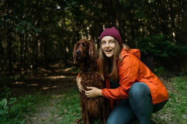 Kadın ve köpek ormanda yürüyüş macerasının tadını çıkarıyorlar. O, çimenlerde otururken köpeği nazikçe tutuyor, ikisi de mutlu görünüyor..