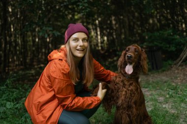 Turuncu ceketli ve bereli neşeli genç bir kadın orman ortamında İrlandalı yerleşimci köpeğiyle poz veriyor. Çiftin etrafı ağaçların arasından süzülen yeşil ve doğal ışıklarla çevrili..