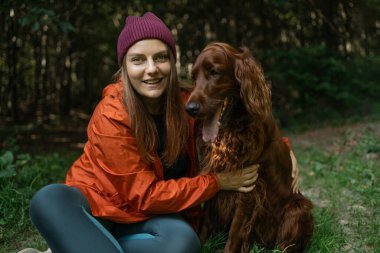 Güler yüzlü kadın ormanda İrlandalı Setter 'ının yanında oturmuş, doğada huzurlu anların tadını çıkarıyor. Evcil hayvan arkadaşlığı ve açık hava eğlencesi temaları için harika..