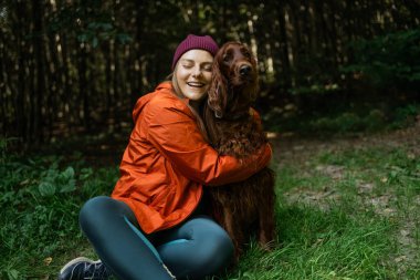 Parlak turuncu ceketli mutlu kadın orman yürüyüşü sırasında köpeğini gözleri kapalı kucaklıyor ve sevgi ve neşe gösteriyor. Bağlanma ve duygusal bağlantıyı temsil eder.
