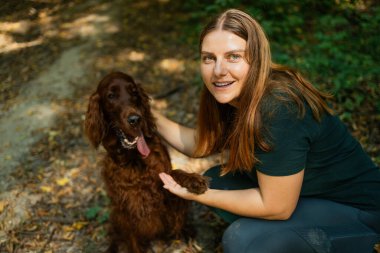 Dost canlısı bir İrlandalı Yerleşimcinin pençesiyle ormanda diz çökmüş gülümseyen bir kadın. Görüntü, doğa yürüyüşü sırasında insanlarla evcil hayvanlar arasındaki bağı gösteren neşeli bir açık hava anı yakalıyor..