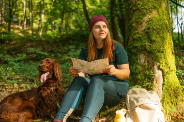 Yürüyüş kıyafetleri içindeki genç bir kadın ormanda bir ağacın altında oturuyor ve yanında köpeğiyle birlikte bir harita okuyor. Yakınlarda bir sırt çantası ve tekrar kullanılabilir su şişesi var. Açık havada barışçıl doğa molası