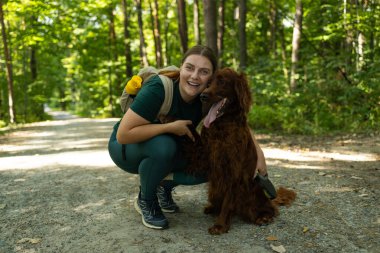 Açık hava kıyafetleri içinde gülümseyen genç bir kadın orman yolunda İrlandalı yerleşimci köpeğinin yanında diz çöker. Neşeli etkileşimleri arkadaşlığı, doğa bağlarını ve vahşi doğadaki macera ruhunu ön plana çıkarıyor..