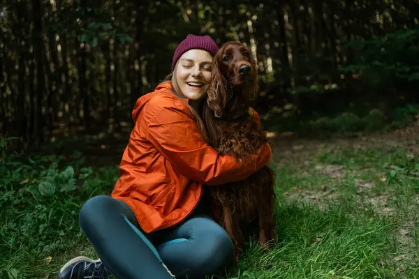 Parlak turuncu ceketli mutlu kadın orman yürüyüşü sırasında köpeğini gözleri kapalı kucaklıyor ve sevgi ve neşe gösteriyor. Bağlanma ve duygusal bağlantıyı temsil eder.