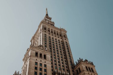 Mavi gökyüzü altında neoklasik mimari detaylara sahip devasa bir gökdelenin alçak açılı görüntüsü. Bina kulesi ve simetrik tasarım, onun büyük ölçekli ve tarihi önemini vurguluyor.