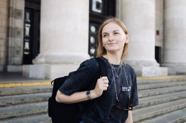 Rahatlamış bir ifadeyle gülümseyen sarışın kadın, günlük kıyafetler, sırt çantası ve boynuna güneş gözlüğü takıyor. Büyük klasik sütunları olan bir binanın önündeki taş basamaklarda durmak