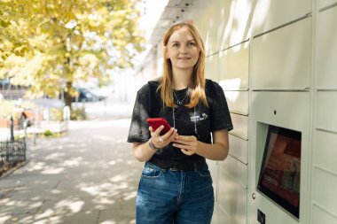 Genç bir kadın güneşli bir günde açık havada bir dolap kullanıyor. Siyah tişörtlü ve kot pantolonlu sıradan bir sarışın kadın elinde akıllı bir telefon, self servis paket terminalinin yanında duruyor. Modern kavram