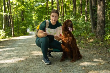 Neşeli bir kadın elinde bir harita tutarken ve orman yolunu işaret ederken İrlandalı Setter köpeğinin yanında diz çöker. Heyecanı, keşfi ve doğada bir evcil hayvanla paylaşılan macerayı yakaladığı an.