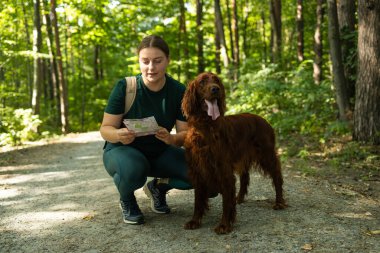 Neşeli bir kadın elinde bir harita tutarken ve orman yolunu işaret ederken İrlandalı Setter köpeğinin yanında diz çöker. Heyecanı, keşfi ve doğada bir evcil hayvanla paylaşılan macerayı yakaladığı an.