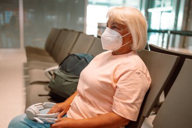 Tıbbi maske takan yaşlı bir kadın havaalanı bekleme odasında oturuyor ve sosyal mesafeyi gözetliyor. Yüksek kalite fotoğraf