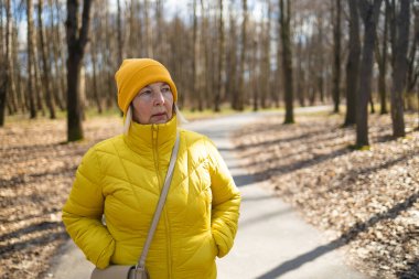 Sarı ceket ve şapka giymiş düşünceli bir kadın, sonbahar ağaçları ve yapraklarla çevrili bir park yolunda duruyor. Huzurlu sahne, değişen mevsimlerin güzelliğini yakalar.