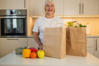 Yaşlı bir kadın taze yiyeceklerle mutfağında duruyor, masada renkli dolmalık biberler ve elmalar da dahil. Etrafı kahverengi poşetlerle çevrili..