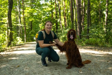 Açık hava kıyafetleri içinde gülümseyen genç bir kadın orman yolunda İrlandalı yerleşimci köpeğinin yanında diz çöker. Neşeli etkileşimleri arkadaşlığı, doğa bağlarını ve vahşi doğadaki macera ruhunu ön plana çıkarıyor..