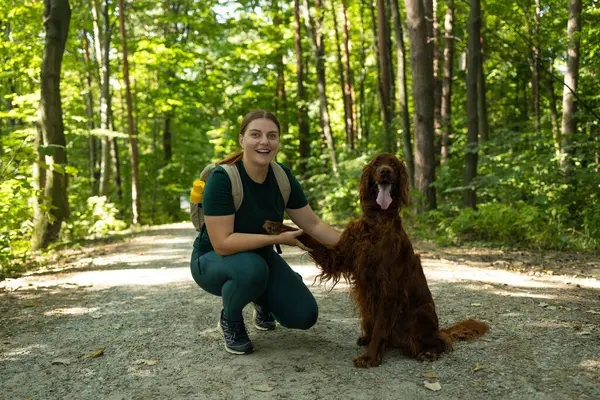 Açık hava kıyafetleri içinde gülümseyen genç bir kadın orman yolunda İrlandalı yerleşimci köpeğinin yanında diz çöker. Neşeli etkileşimleri arkadaşlığı, doğa bağlarını ve vahşi doğadaki macera ruhunu ön plana çıkarıyor..