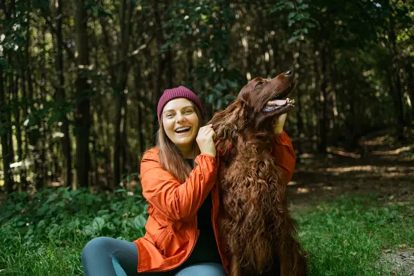 Ormanda köpeğiyle birlikte heyecanlı bir kadın yürüyüşçü, çömelmiş ve sarılıyor. Eğlence, açık hava aktivitesi ve evcil hayvanlarla arkadaşlık vurgulanıyor.