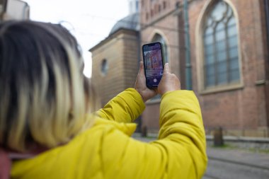 Sarı ceketli bir turist akıllı telefonuyla tarihi bir binayı yakalıyor. Görüntü, teknolojiyle kültürel keşifleri birleştiren popüler bir seyahat aktivitesi sergiliyor..