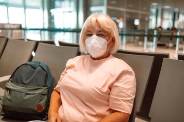 Tıbbi maske takan yaşlı bir kadın havaalanı bekleme odasında oturuyor ve sosyal mesafeyi gözetliyor. Yüksek kalite fotoğraf