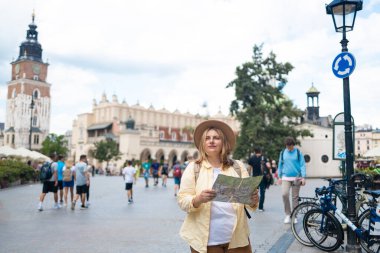 Sarı ceketli ve şapkalı bir kadın canlı bir şehir meydanında elinde haritayla bölgeyi dolaşmaya çalışıyor. Şehir mimarisi ve bisikletçileriyle turist gibi görünüyor.