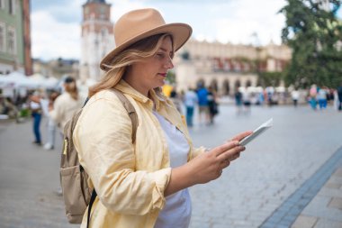 Sarı ceketli ve şapkalı bir kadın canlı bir şehir meydanında elinde haritayla bölgeyi dolaşmaya çalışıyor. Şehir mimarisi ve bisikletçileriyle turist gibi görünüyor.