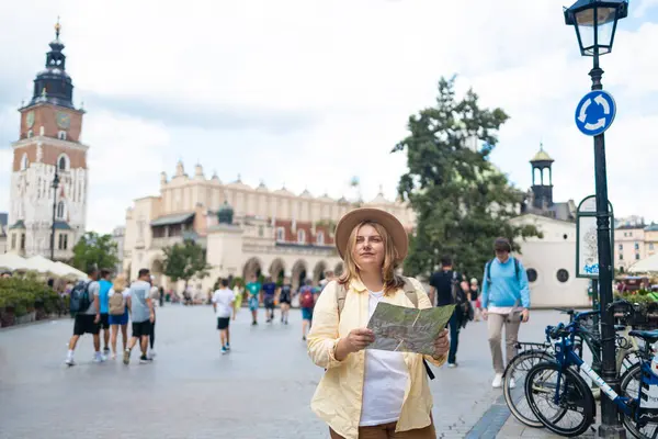Sarı ceketli ve şapkalı bir kadın canlı bir şehir meydanında elinde haritayla bölgeyi dolaşmaya çalışıyor. Şehir mimarisi ve bisikletçileriyle turist gibi görünüyor.