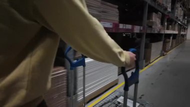 Sarı kazaklı bir insan mobilya deposundaki koridorda düz yatak arabasını itiyor. Raflar ve paketlenmiş mallarla çevrelenmiş olan bu resim, kendi kendine hizmet veren alışveriş deneyimlerini büyük ölçüde yansıtıyor.