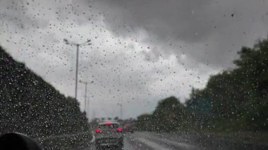 Yağmur damlaları, araçlar karanlık ve bulutlu bir otoyolda ilerlerken ön camı kapatır. Fırtınalı atmosfer, bulanık ışıklar ve ıslak yol şiddetli yağmur ve zorlu hava koşullarında yolculuk havasını yakalar.