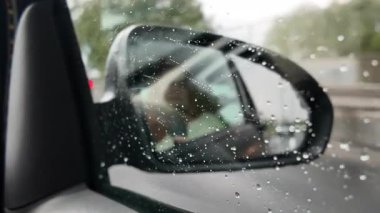Yağmurlu bir günde arabanın yan aynasına su damlacıklarıyla yakın çekim. Islak cam bulanık bir otoyol sahnesini yansıtıyor. Kötü havada araba sürme atmosferini ve yağmurun görsel dokusunu yakalıyor.