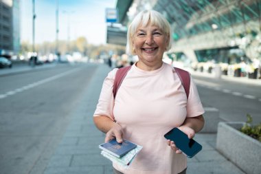 Pasaportunu ve akıllı telefonunu tutan yaşlı bir kadın havaalanı terminalinin dışında mutlu bir şekilde gülümsüyor. Yolculuğu için gerekli belgeler ve seyahat teknolojisiyle hazırlandı.