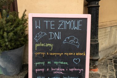Üzerinde el yazısı Lehçe yazı olan büyüleyici bir kafe tahtası her daim yeşil bir bitkinin yanındaki kaldırımlı bir sokakta duruyor. Kurul, mevsimlik rahat bir atmosfer yaratarak kış spesiyallerini teşvik ediyor.