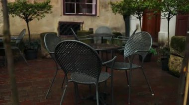 Açık bir kafede boş metal sandalyeler ve masalar yağmurlu havada ıslanmış bir şekilde otururlar. Saksı çiçekleri, arka planda iki yaya yürürken, oturma alanının yanındaki taş ekicisi sıralanır..