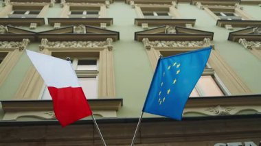 Süslü tarihi bir binanın önünde Polands ulusal bayrağı ve Avrupa Birliği bayrağı dalgalanıyor. Görüntü, bir kentsel ortamda ulusal ve Avrupa kimliğinin sembolik bir temsilini yakalıyor..