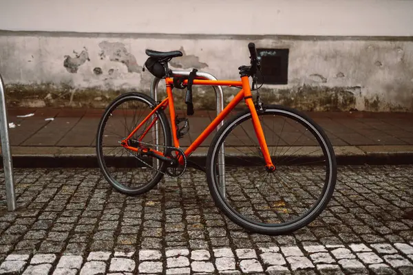 Parlak turuncu bisiklet parkedilmiş ve kaldırım taşı caddesindeki metal bisiklet parkına sabitlenmiş. Arkasında, beyaz çerçeveli camları olan tarihi bir bej bina duruyor. Sakin, kentsel bir Avrupa ortamını çağrıştırıyor.