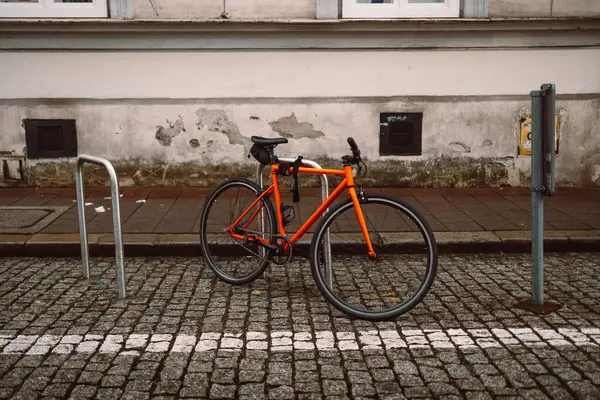 Parlak turuncu bisiklet parkedilmiş ve kaldırım taşı caddesindeki metal bisiklet parkına sabitlenmiş. Arkasında, beyaz çerçeveli camları olan tarihi bir bej bina duruyor. Sakin, kentsel bir Avrupa ortamını çağrıştırıyor.