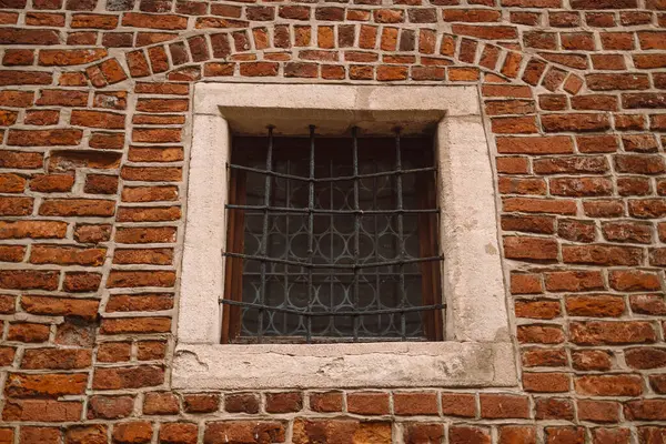 Küçük, kare bir pencere, demir parmaklıklar ve eski kırmızı tuğla bir duvar. Işık taşıyla çerçevelenen pencere, tarihi veya güçlendirilmiş bir mimari tarzını anımsatarak, desen ve karakteri de ekliyor