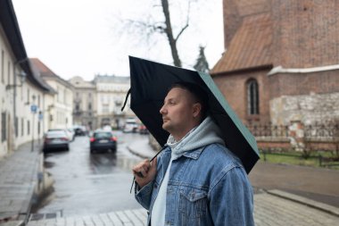 Kot ceketli ve kapüşonlu bir adam yağmurlu bir Avrupa caddesinde büyük siyah bir şemsiye tutarak yürüyor. Arka planda tarihi mimari ve ıslak kaldırım taşları yer alıyor, karamsar ve bulutlu bir görüntü yakalıyorlar.