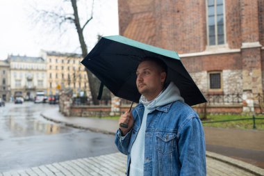 Kot ceketli ve kapüşonlu bir adam yağmurlu bir Avrupa caddesinde büyük siyah bir şemsiye tutarak yürüyor. Arka planda tarihi mimari ve ıslak kaldırım taşları yer alıyor, karamsar ve bulutlu bir görüntü yakalıyorlar.